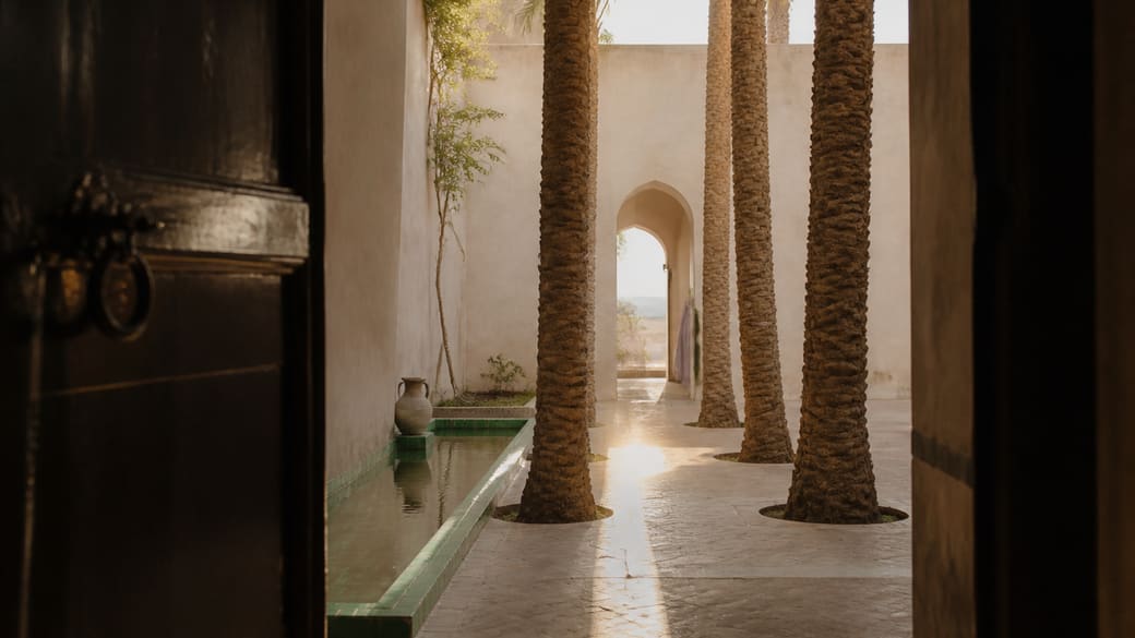 Vue conceptuelle au lever du jour depuis une porte sombre vers un patio ivoire structuré par quatre palmiers